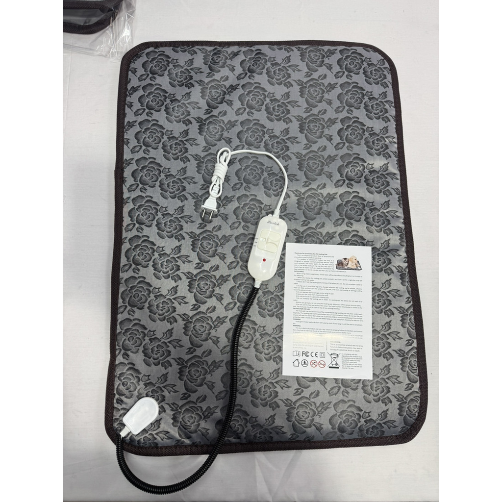An electric heated mat with a gray and black floral pattern, shown with its white power cord, controller, and an instruction manual on a white surface.